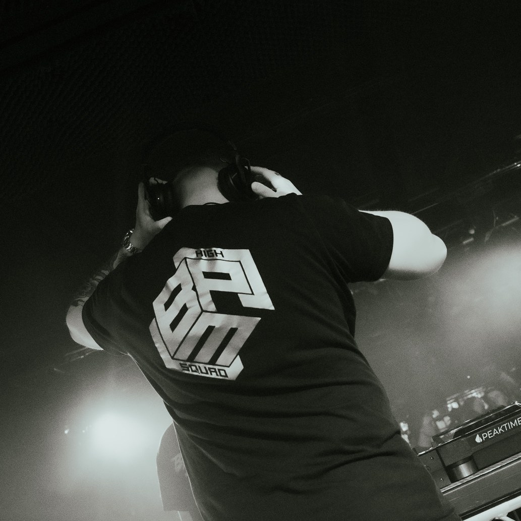 Black and white shot of a DJ in a High BPM Squad shirt behind the decks, smoke and stage lights